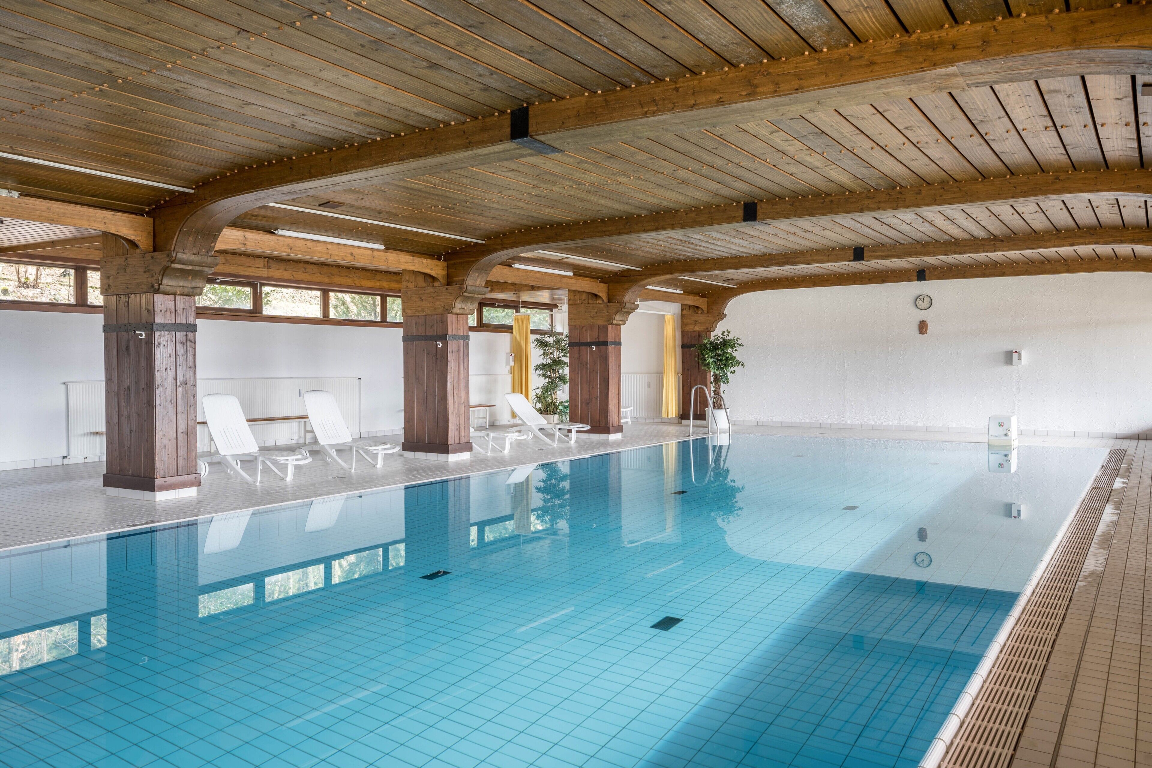 En indendørs swimmingpool med træbjælker, hvide fliserede vægge og hvide liggestole.