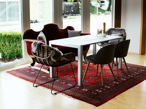 Een eetruimte met een witte tafel, rode en zwarte stoelen, een kamerbreed tapijt en uitzicht op de omgeving door grote ramen.