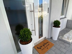 A modern entrance with a glass door, two potted plants, a doormat with text, and a stone sculpture on a gray bench. - Apartment 'Schwarzwaldzauber' with Mountain View, Private Terrace and Wi-Fi (Sankt Blasien)