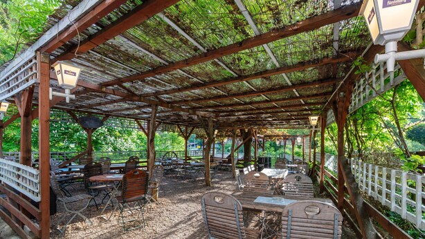 En udendørs overdækket terrasse med borde og stole, et træbeklædt pergola og et hvidt hegn.