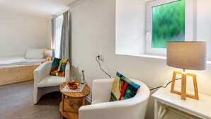 Een hotelkamer met een bed, een witte fauteuil, een klein houten tafeltje, een lamp en een raam met uitzicht op groen.