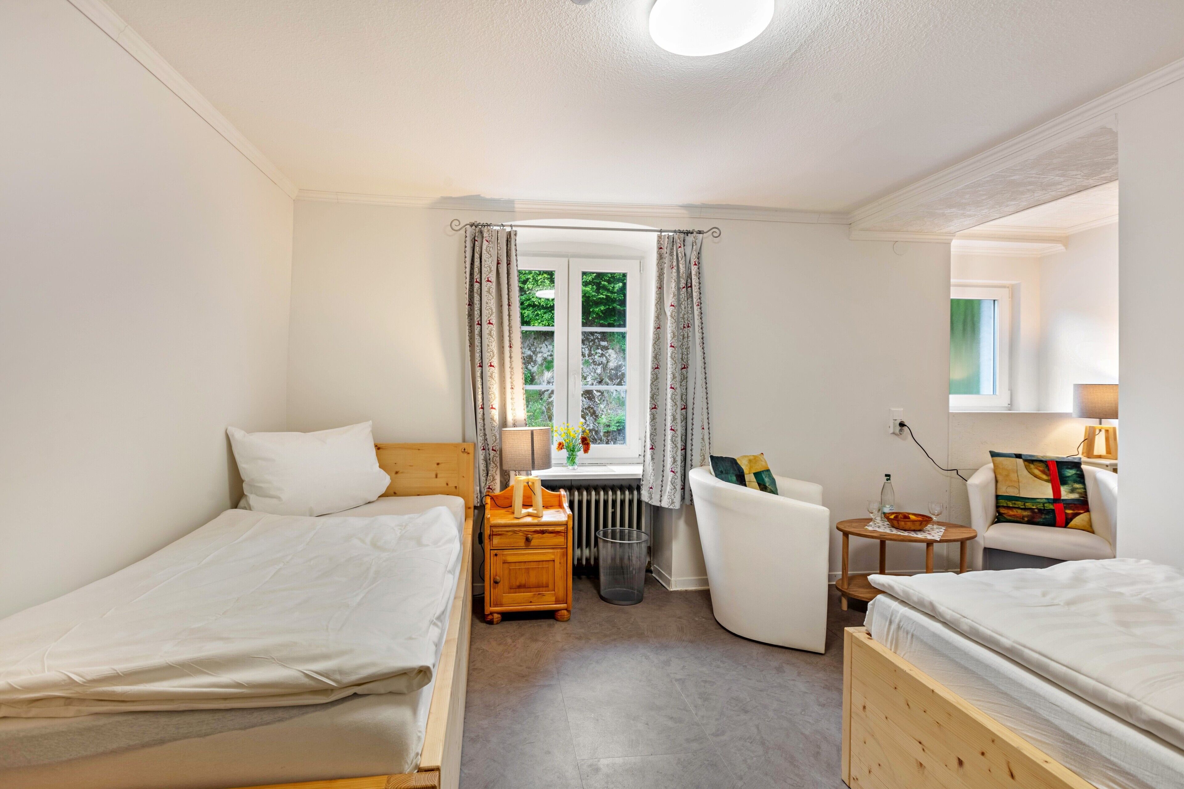 Ett rum med två sängar, ett fönster med gardiner, ett element, ett litet bord med en lampa och en stol.
