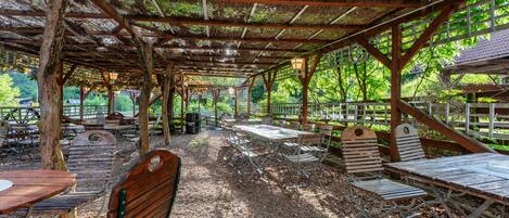An outdoor covered patio with tables and benches, surrounded by trees.