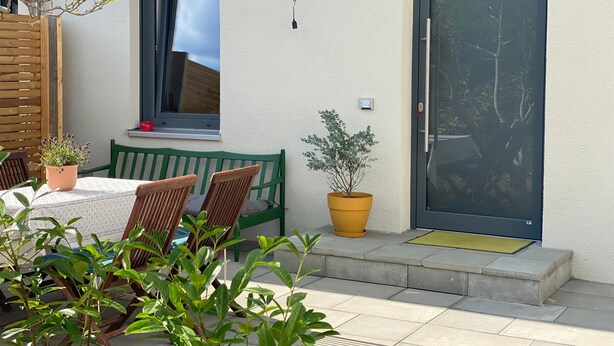 Une terrasse avec une table et des chaises, un pot de fleurs, une fenêtre et une porte.