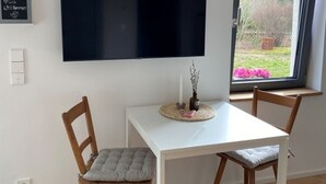 Une pièce avec une table blanche, deux chaises en bois, un téléviseur à écran plat noir fixé au mur, une petite plante en pot sur la table et une fenêtre donnant sur un paysage verdoyant.