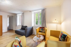 Una habitación con un sofá gris, sillones amarillos, una mesita redonda con una botella y vasos, una lámpara y una ventana con cortinas.