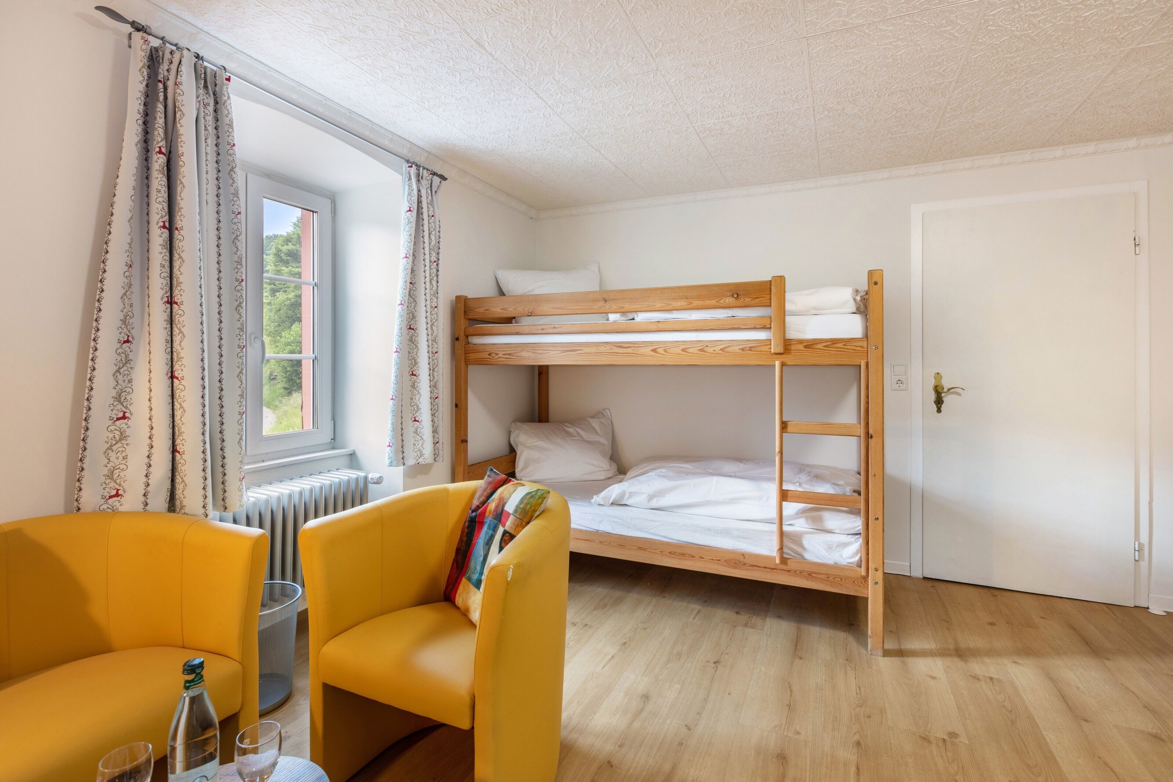 Una habitación con literas, un sillón amarillo, un radiador y una ventana con cortinas.
