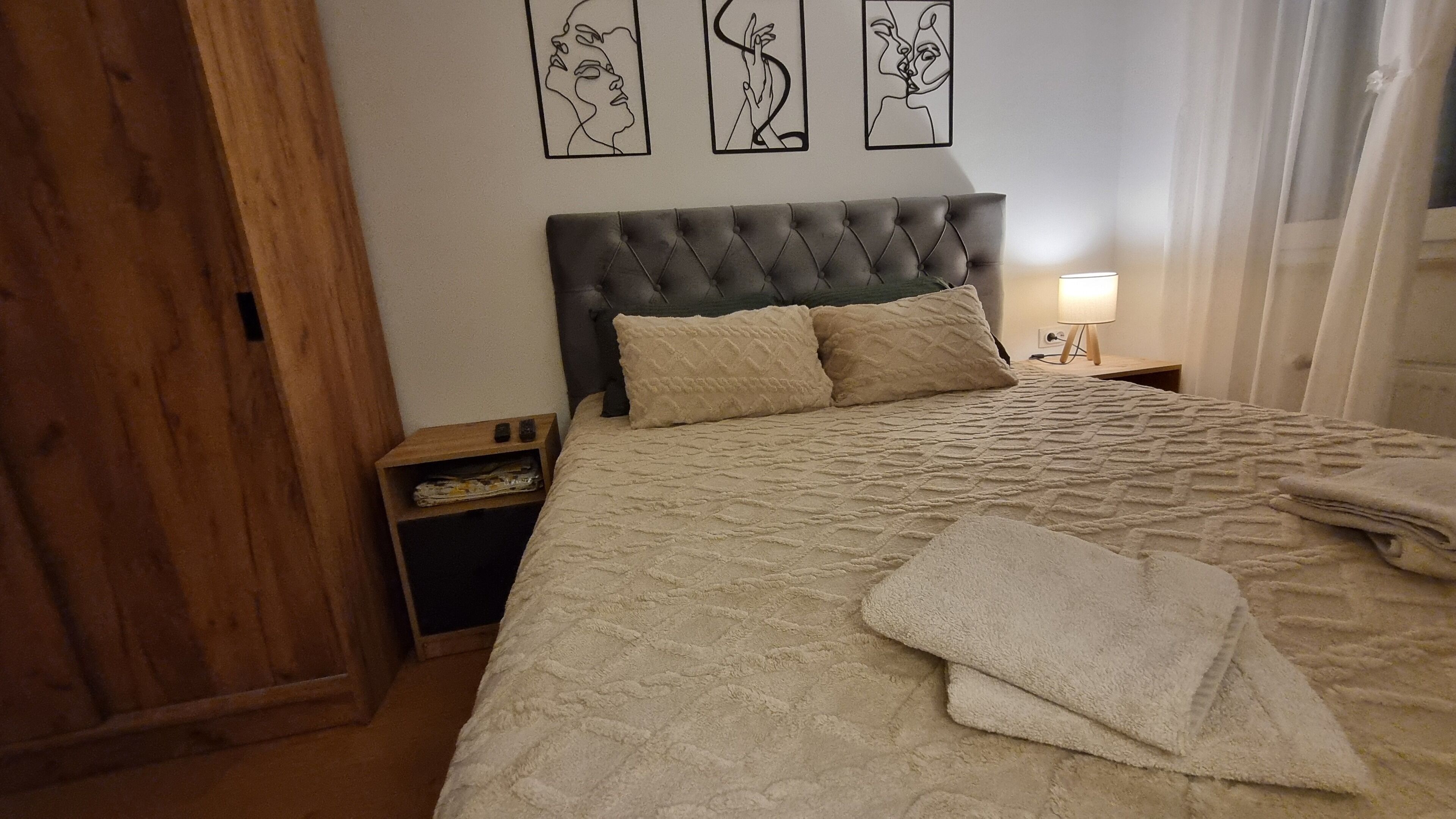 Ein Schlafzimmer mit einem gepolsterten Kopfteil, einem Bett mit einer strukturierten Decke, zwei Kissen, einem Nachttisch mit einer Lampe und einem Kunstwerk an der Wand.