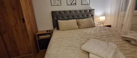 Ein Schlafzimmer mit einem gepolsterten Kopfteil, einem Bett mit einer strukturierten Decke, zwei Kissen, einem Nachttisch mit einer Lampe und einem Kunstwerk an der Wand.