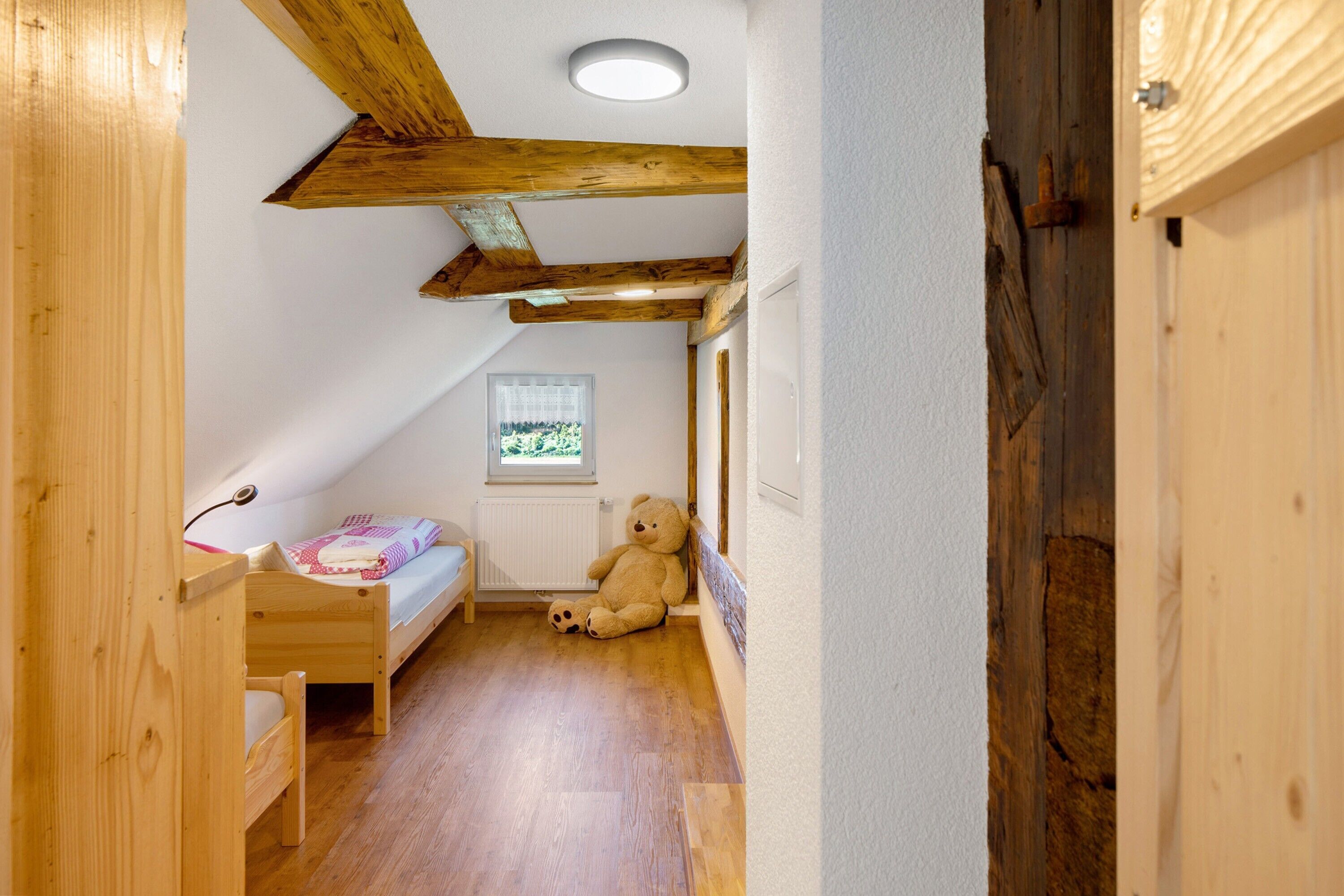 Une petite pièce chaleureuse et accueillante, avec un lit superposé en bois, un ours en peluche, une fenêtre donnant sur un beau paysage et des poutres apparentes en bois au plafond.