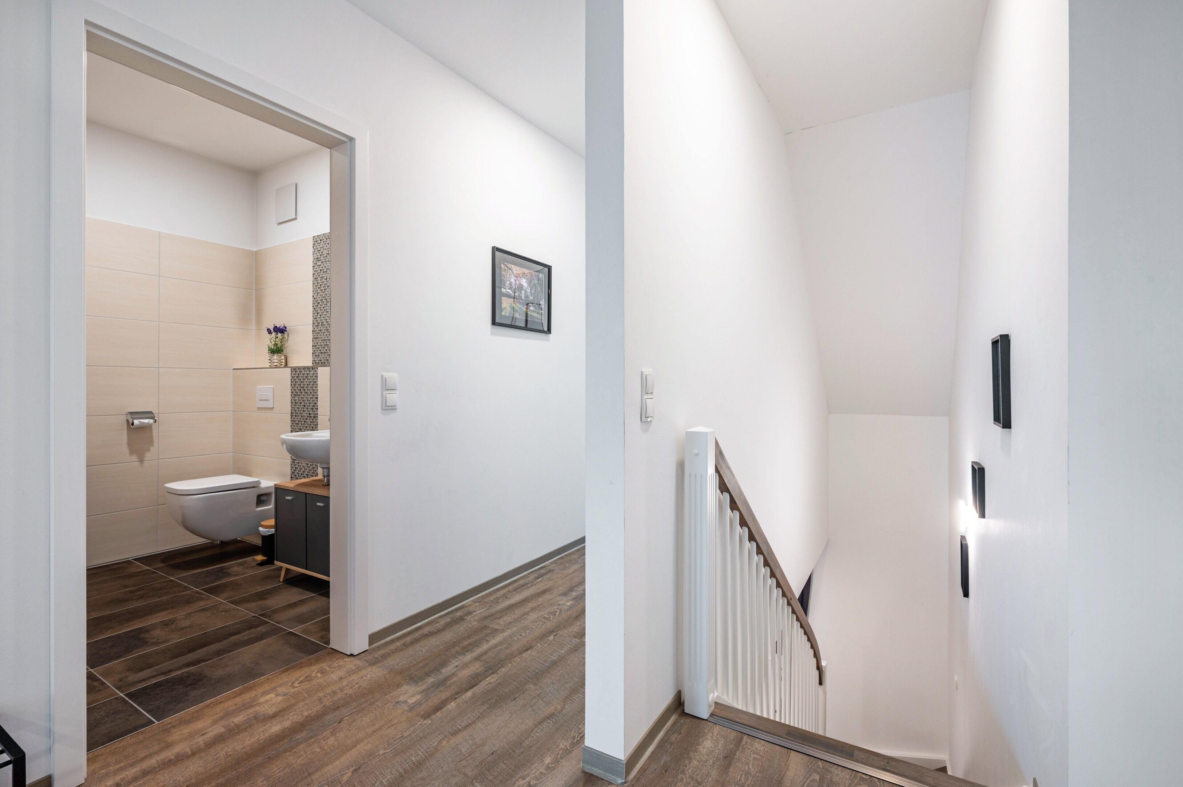 A hallway with a wooden floor, a white wall, a white radiator, and a staircase with a white railing.