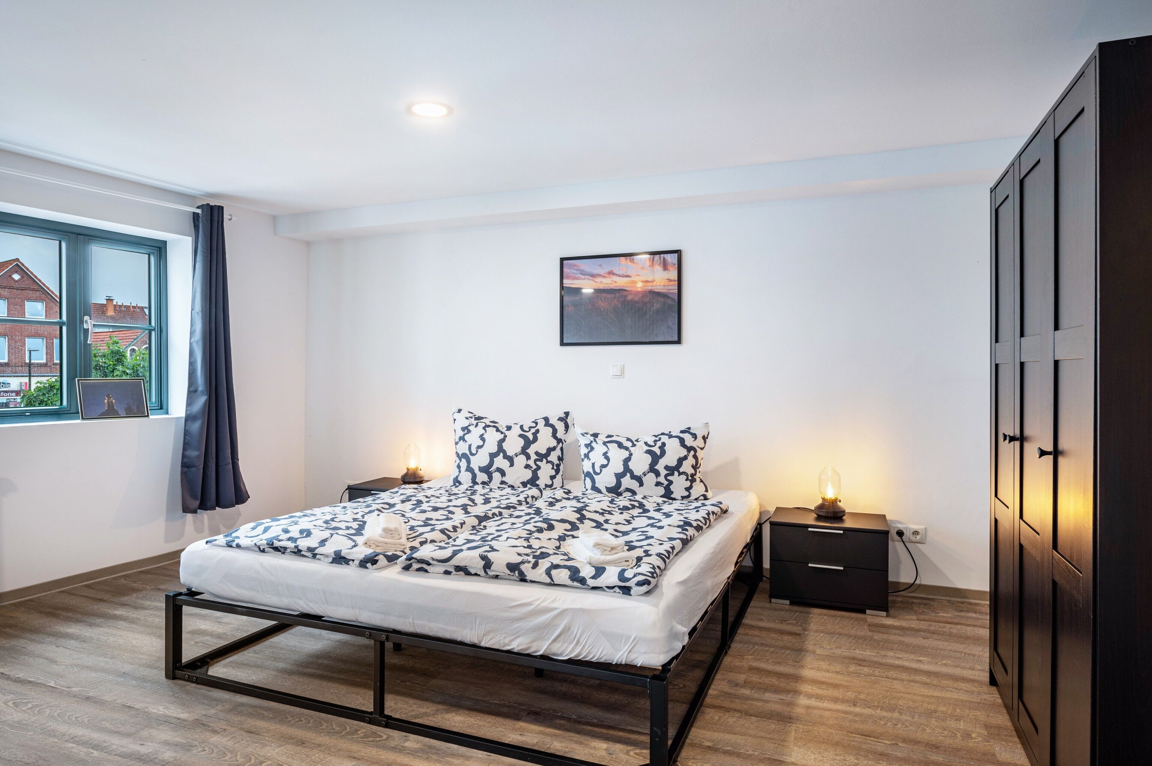 Un dormitorio con cama, mesita de noche, un cuadro en la pared y ventana con cortinas.