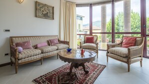 Ein Wohnzimmer mit einer Couch, Sesseln, einem Couchtisch und einem großen Fenster mit Blick auf Grünflächen und Gebäude.