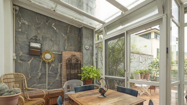 Ein Wintergarten mit Steinwand, einem Holztisch und Korbstühlen.