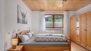 Une chambre à coucher avec un lit en bois, une table de chevet, une lampe, une fenêtre donnant sur un lac et une armoire en bois.