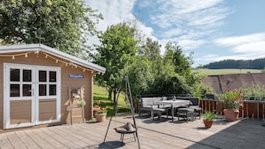 Une terrasse en bois avec un fauteuil suspendu, un petit bâtiment portant l’inscription « Königsallee » et du mobilier d’extérieur.