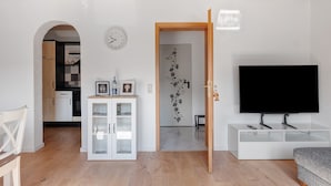 Un salon moderne avec un téléviseur, des meubles blancs, un plafond en bois et une cuisine en arrière-plan.