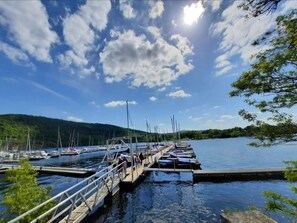 Un puerto deportivo con barcos amarrados, un muelle de madera y una colina boscosa al fondo.