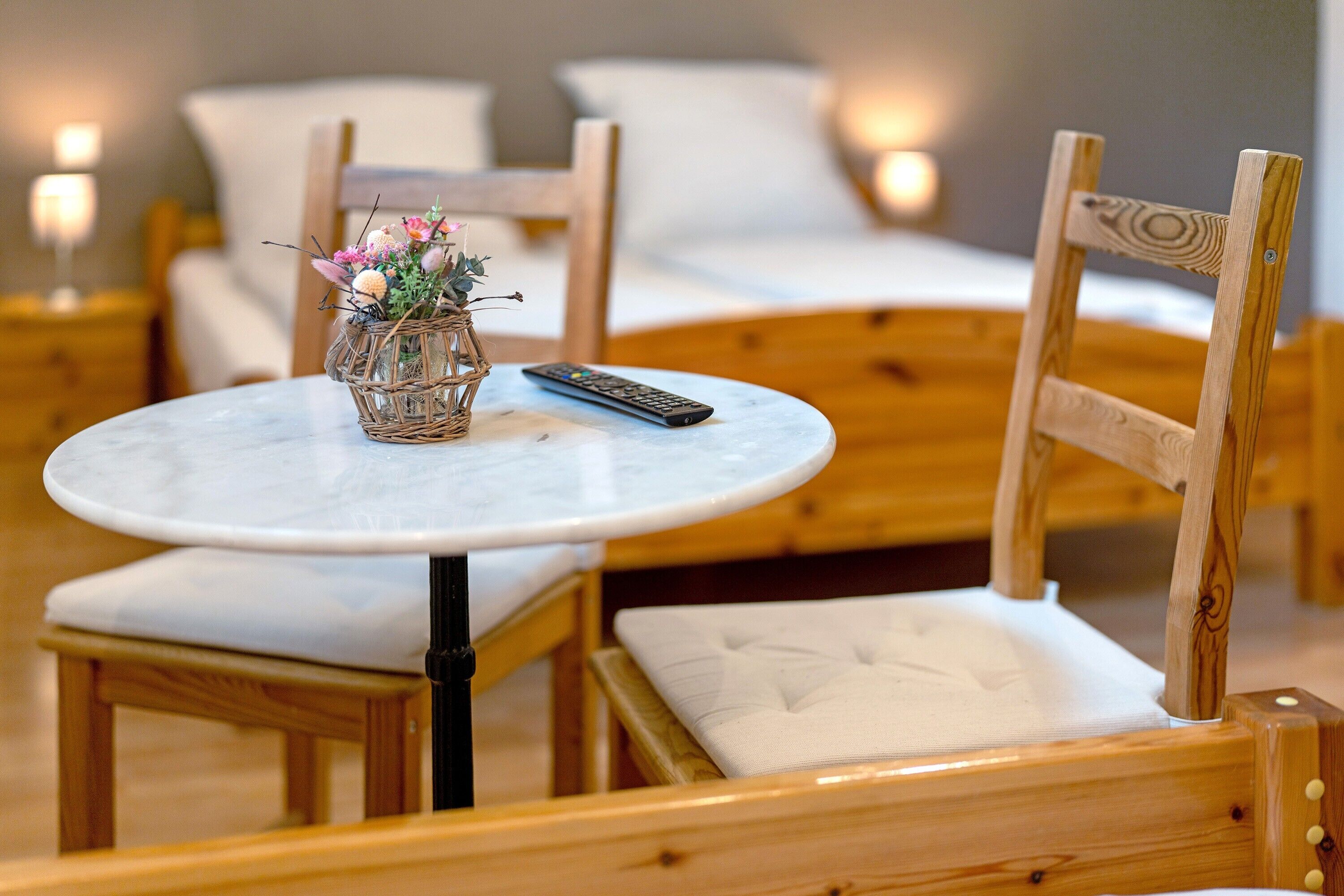 A wooden chair with a cushioned seat and backrest, a small round table with a remote control and a glass vase with flowers, and a bed with white bedding in the background.