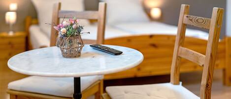 A wooden chair with a cushioned seat and backrest, a small round table with a remote control and a glass vase with flowers, and a bed with white bedding in the background.