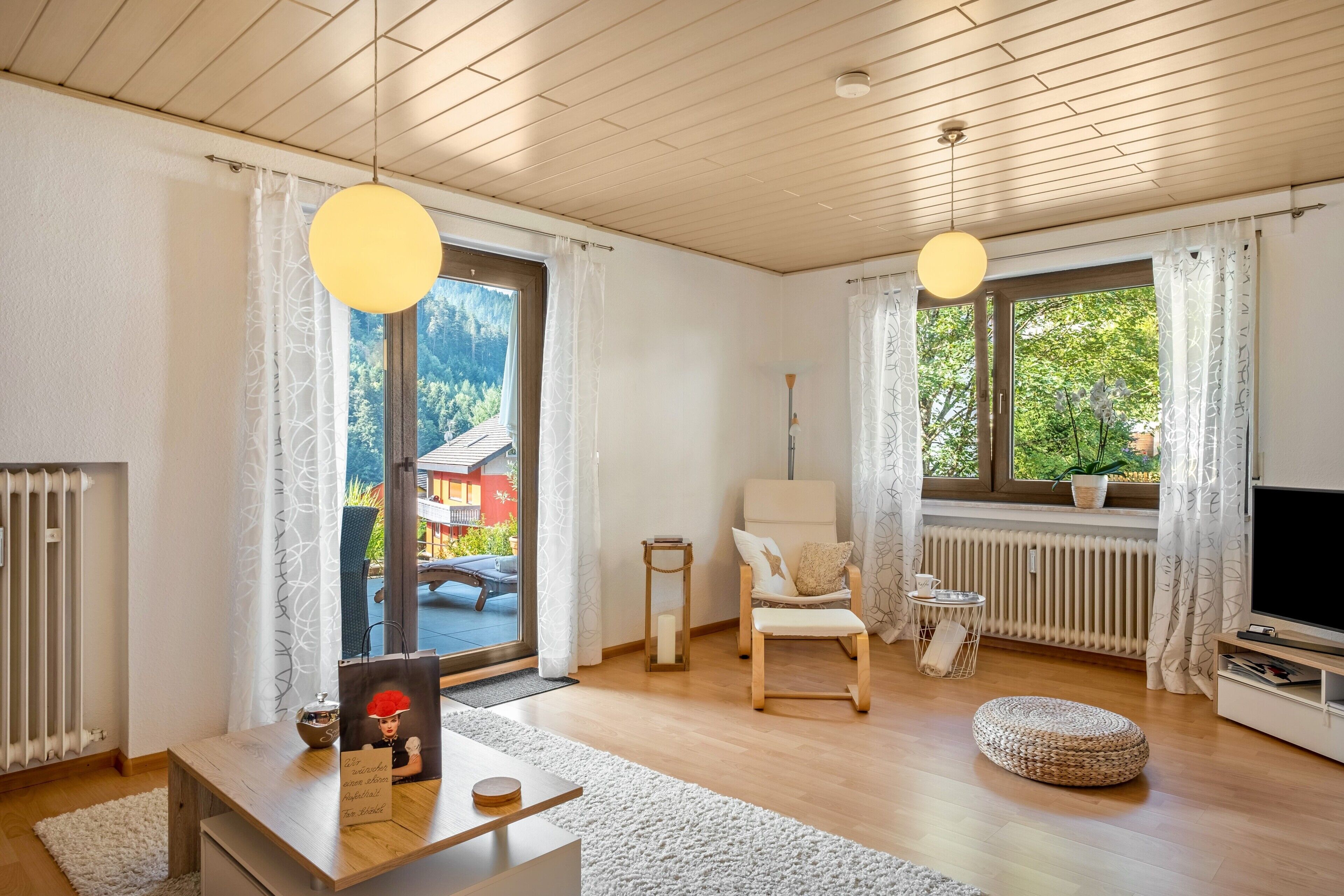 Una sala de estar moderna con piso de madera, un sofá, una silla, un televisor y un ventanal con vistas a una casa y árboles.