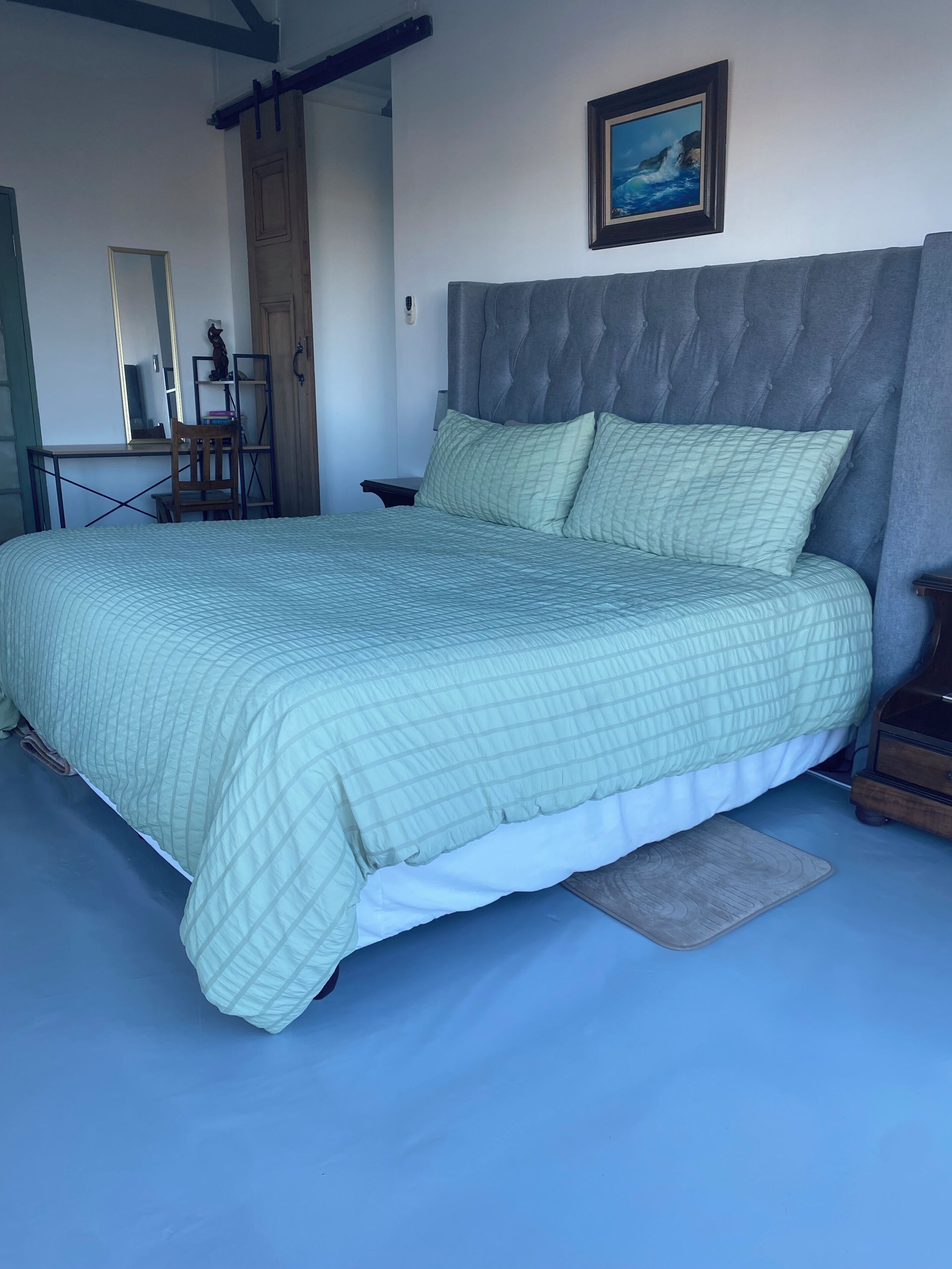 A neatly made bed with a quilted bedspread, a wooden chair, a nightstand, a framed picture on the wall, and a door with a wooden frame.