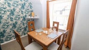 A dining area with a wooden table set for four, surrounded by chairs with cushioned seats. A window with curtains is visible in the background. - Medical Hall (Cemaes Bay)