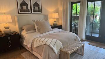 A bedroom with a bed, two bedside tables with lamps, a bench, a large window with curtains, and two framed pictures on the wall.