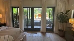 Ein Schlafzimmer mit Bett, einer Sitzbank, einer Lampe, einem Schminktisch und Blick auf eine Terrasse mit Stühlen und Pflanzen.