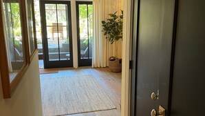 Ein Flur, der zu einer dunklen Holztür führt, mit einem großen Topfpflanzen und Blick durch Glastüren auf eine Außenpatio mit Tisch und Stühlen.