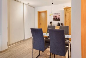 Quarto de hotel com chão de madeira, uma mesa de jantar com cadeiras, um armário de madeira, uma porta e um candeeiro suspenso.