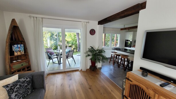 A living room with a sofa, a coffee table, a TV, a wooden bookshelf, a dining area with chairs, a potted plant, and a sliding glass door leading to an outdoor patio. - Apartment 'Schwarzwald-Traum' with Mountain View, Private Terrace and Wi-Fi (Haslach im Kinzigtal)