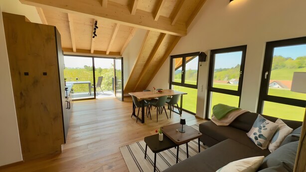 A spacious loft with a high ceiling, a dining area with a table and chairs, a sofa with cushions, and large windows offering a view of the outdoors. - Holiday Home 'Steinackerhof - 2' with Mountain View, Shared Garden and Wi-Fi (Dachsberg (Südschwarzwald))