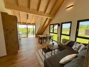 A spacious loft with a high ceiling, a dining area with a table and chairs, a sofa with cushions, and large windows offering a view of the outdoors.
