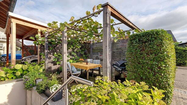 Una pérgola de madera con una mesa y sillas debajo, rodeada de plantas diversas y un seto.
