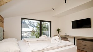 A modern hotel room with a large bed, a desk, a flat-screen TV, and a view of a snowy mountain through a large window. - Apartment in Berwang with Ski-In/Ski-Out (Berwang)