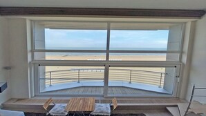 Habitación con un ventanal grande con vista a la playa. La habitación tiene un balcón curvo con barandilla metálica y una pequeña mesa con dos sillas.