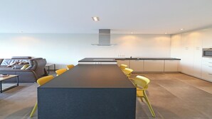 Une cuisine moderne avec une longue table et des chaises jaunes, un canapé gris et une grande fenêtre donnant sur une étendue d’eau.