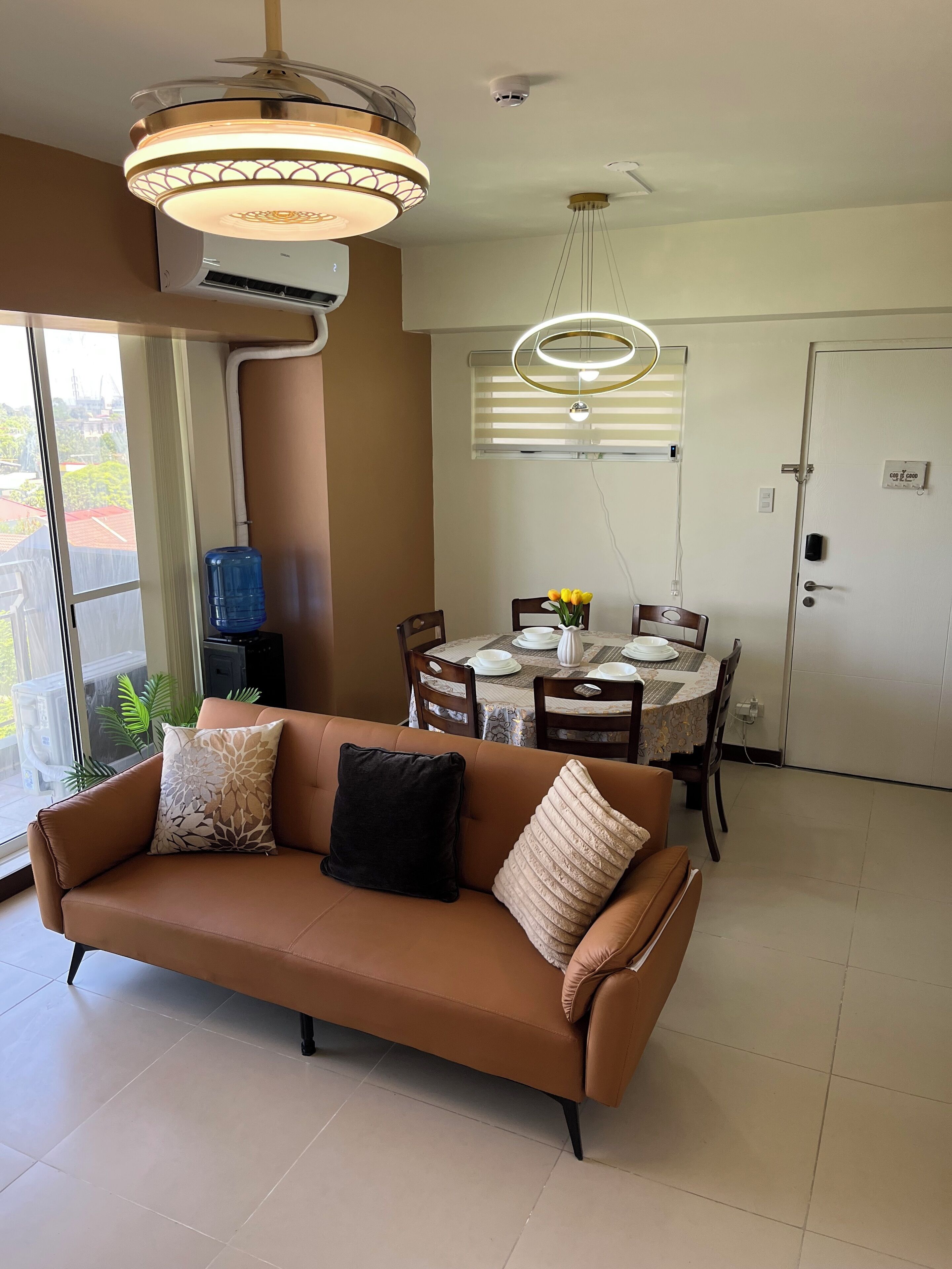 A living room with a brown sofa, a dining table set with plates and a vase of flowers, and a ceiling light fixture.