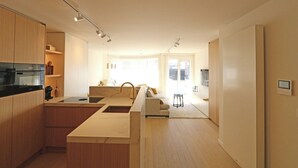 Eine moderne Küche mit Holzausbau, einer zentralen Kochinsel und integrierten Geräten. Ein Essbereich mit einer Couch und einem kleinen Tisch lädt zum Verweilen ein. Ein großes Fenster sorgt für natürliches Licht.