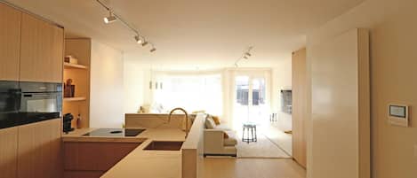 A modern kitchen with wooden cabinets, a central island, and built-in appliances. There is a dining area with a sofa and a small table. The room has a large window providing natural light.
