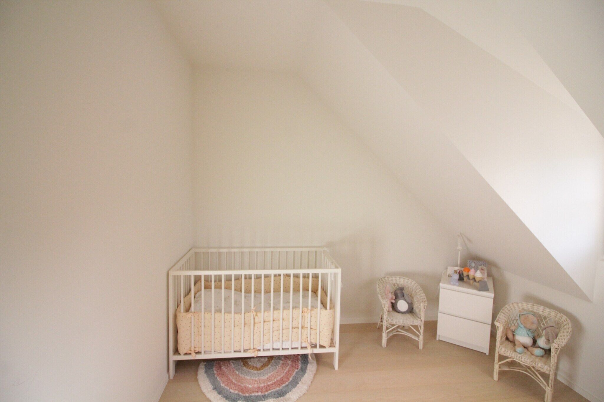 Ein Zimmer mit hohen Decken, einem weißen Babybett, einem Korbstuhl und einem kleinen Nachttisch.