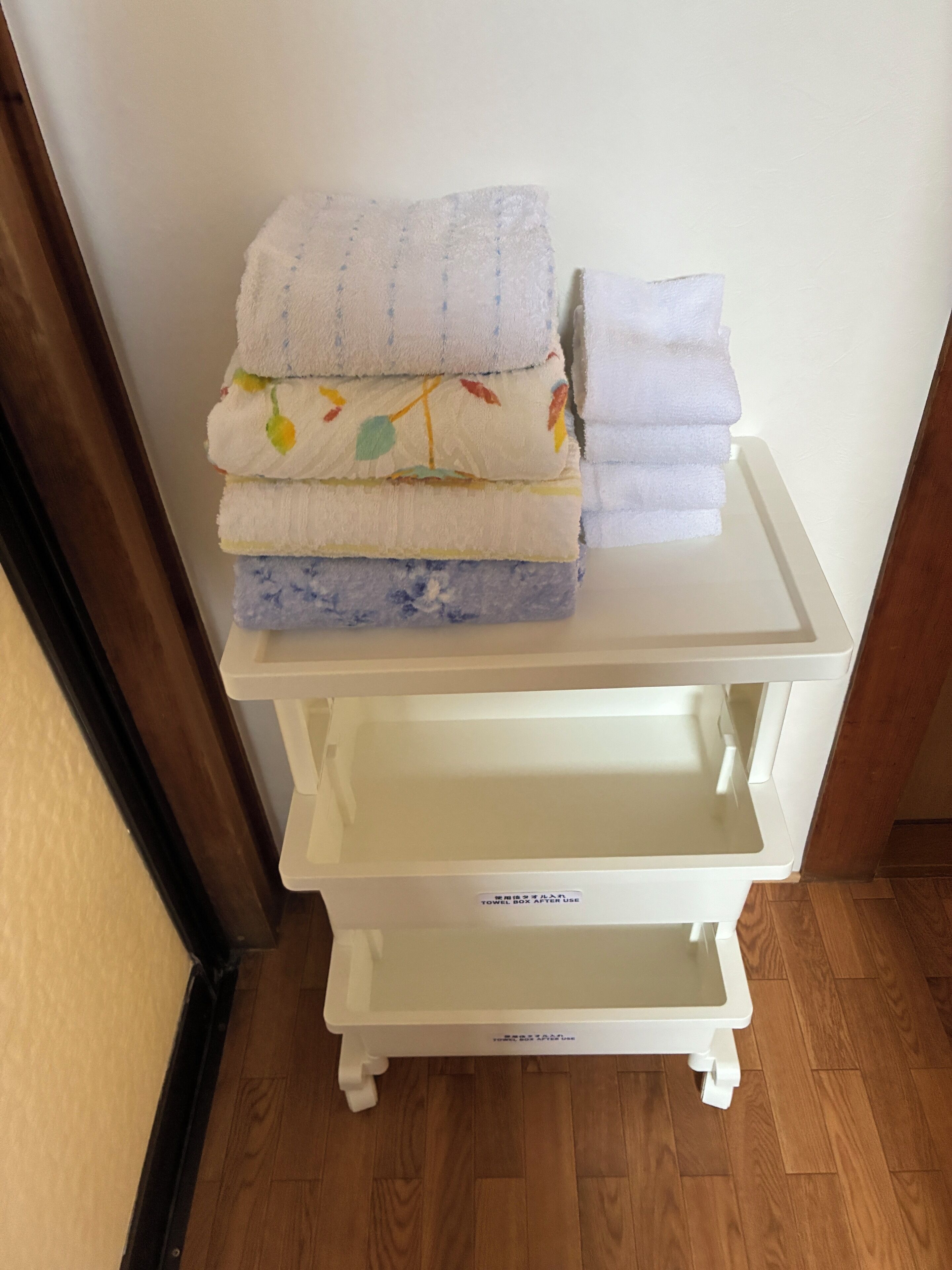 A white trolley with a label 'TOWEL BOX' placed on a wooden floor against a wall.