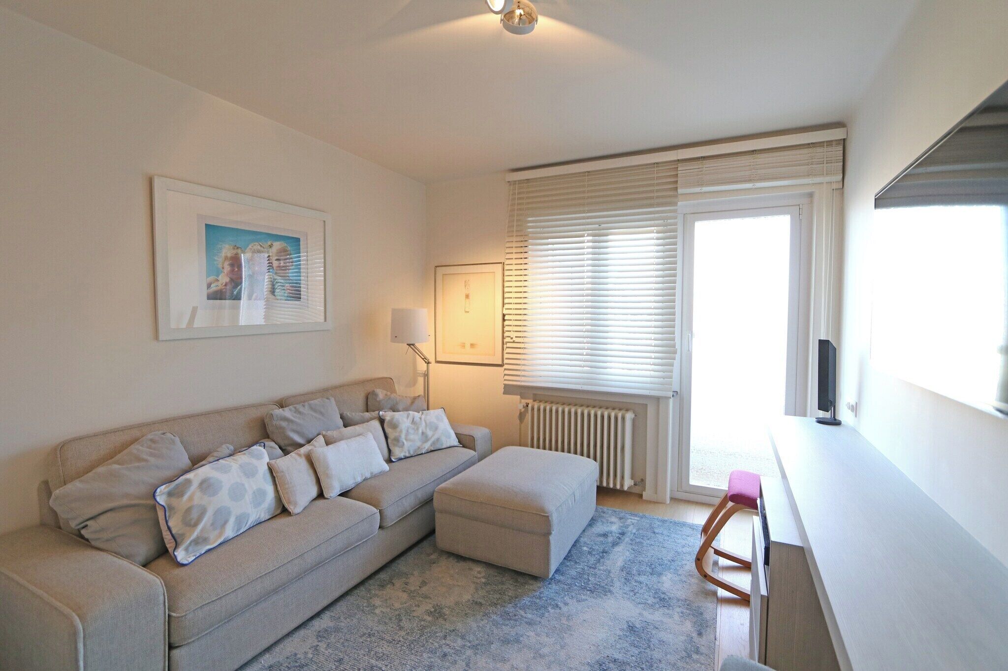 Un salon avec un canapé, un pouf et une petite table. On y trouve un radiateur, une fenêtre avec des stores et un tableau encadré accroché au mur.