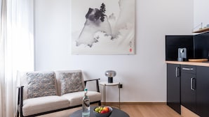 Un salon moderne comprenant un canapé, une table basse avec un saladier de fruits et une bouteille, une petite table d’appoint avec une lampe, et un coin cuisine avec des placards et un plan de travail.