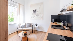 Un salon moderne comprenant un canapé, une petite table ronde avec un saladier de fruits, un tableau au mur et un coin cuisine avec un plan de travail et des appareils électroménagers.