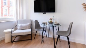 Una sala de estar moderna con un televisor de pantalla plana montado en la pared, una mesa redonda de cristal con dos sillas, un sofá blanco con un cojín estampado y una ventana con cortinas.