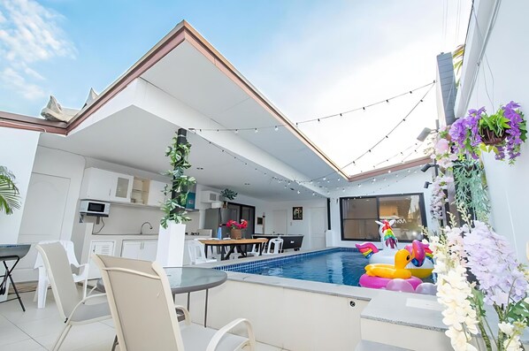 A modern outdoor pool area with a dining setup, a kitchen in the background, and decorative plants.