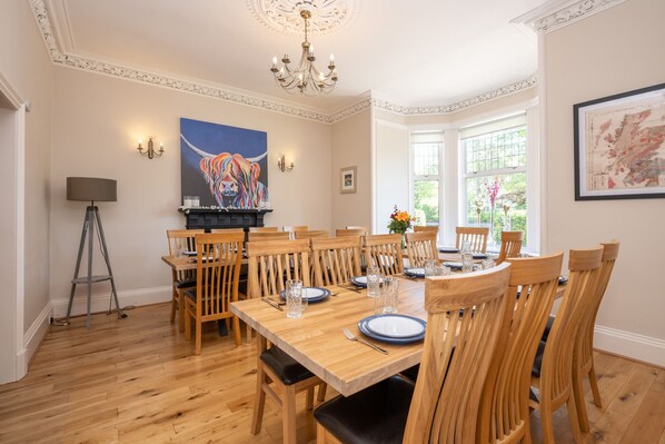 A dining room with a wooden table set for six, surrounded by wooden chairs. There is a chandelier above the table, a floor lamp to the left, and a painting on the wall.