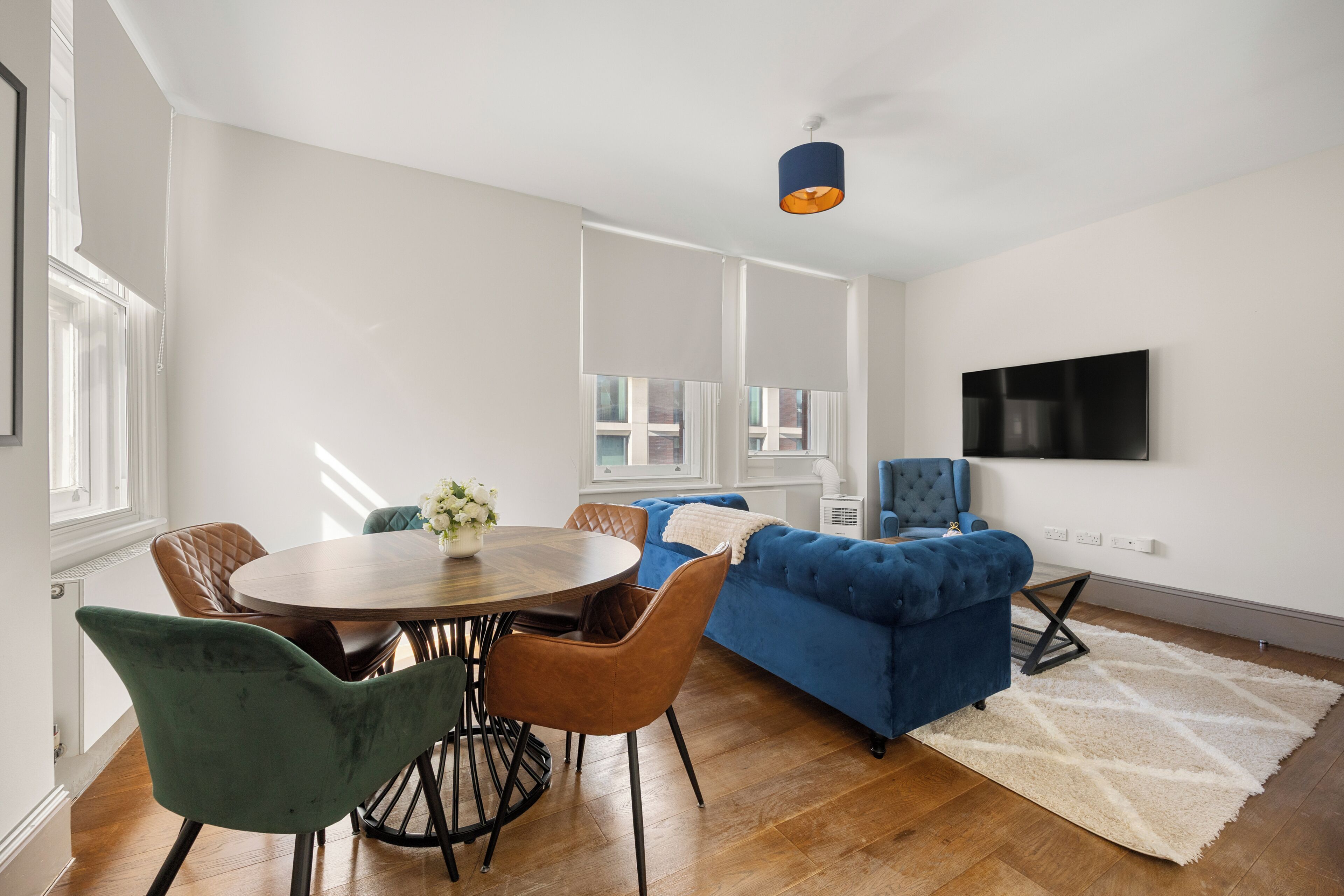 A modern living room with a round dining table, blue sofa, and a flat-screen TV mounted on the wall.